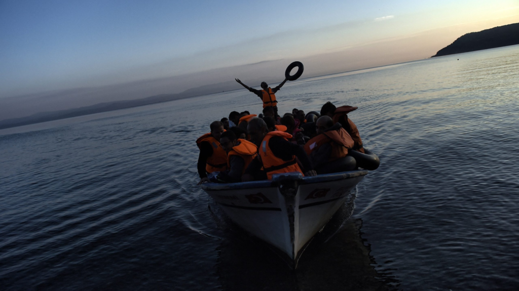 A photo of people in orange life vests seated on a small boat in the sea. One person at the far end of the boat stands with arms raised to the sky and a life preserver in one hand.