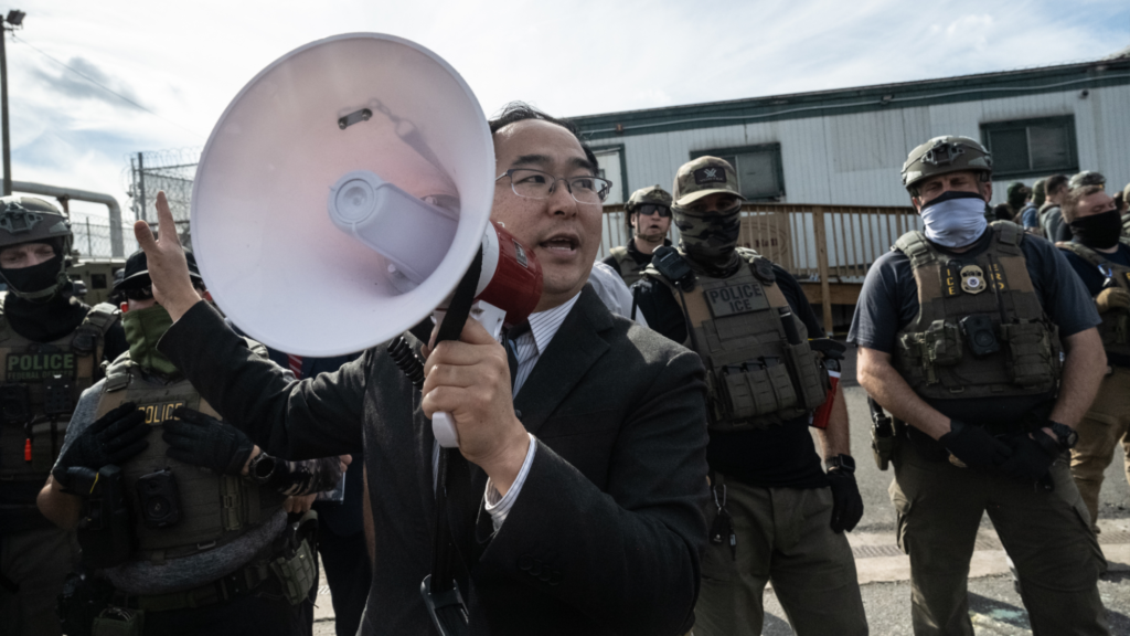 Andy Kim is wearing a suit and is speaking into a megaphone that he is holding in his left hand. Behind him are several armed officers with tactical gear. They are all standing outside an immigration detention center.