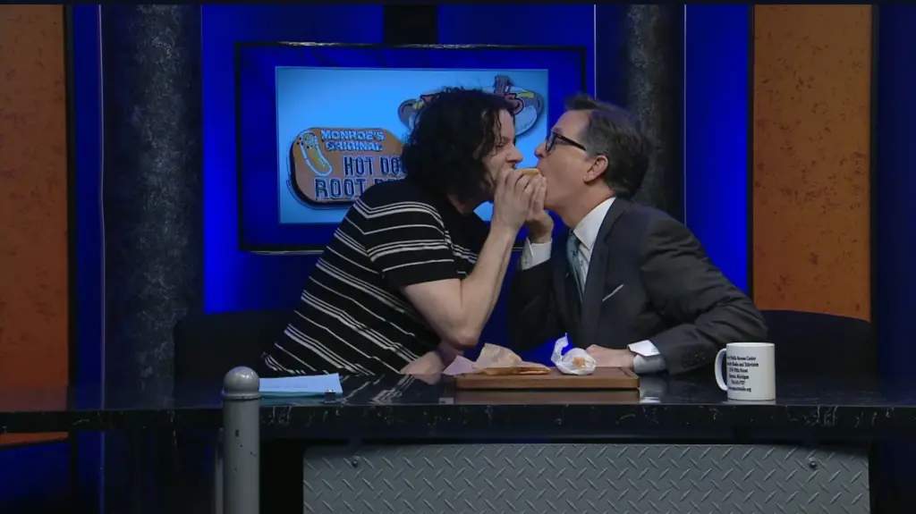 Jack White and Stephen Colbert eat a hot dog together behind a desk.