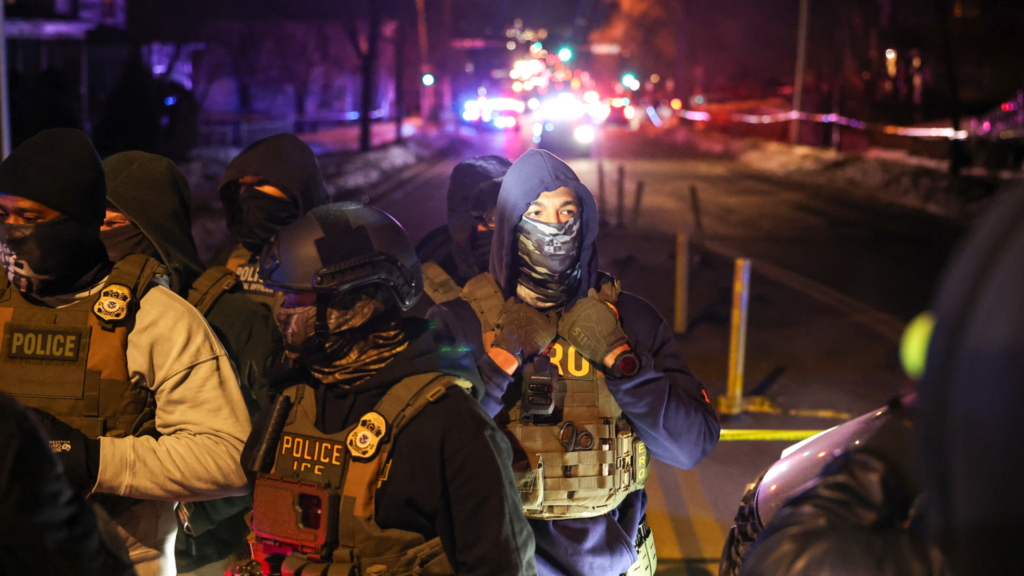 A crowd of about half a dozen officers are standing on a street in the front of a photo as cars are seen in the background. The officers are wearing tactical vests.