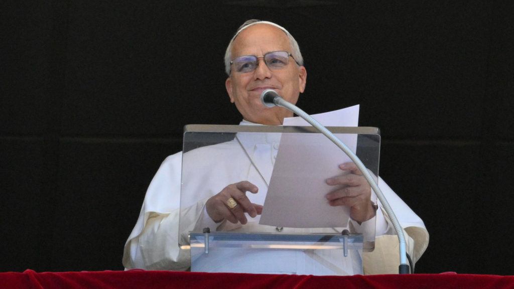 Pope Leo holds paper in his left hand as he speaks into a microphone on a stand from a window. He is wearing white robes, a ring on the fourth finger of his right hand, and glasses.
