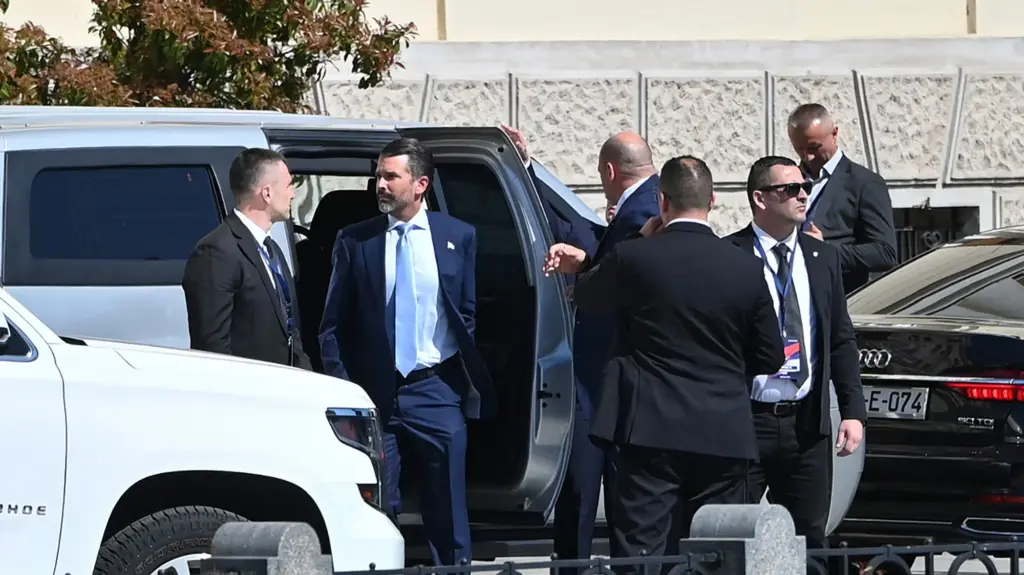 Donald Trump Jr. clad in a dark suit and lavender tie exits a car, surrounded by other men in suits, some of whom are clearly bodyguards.
