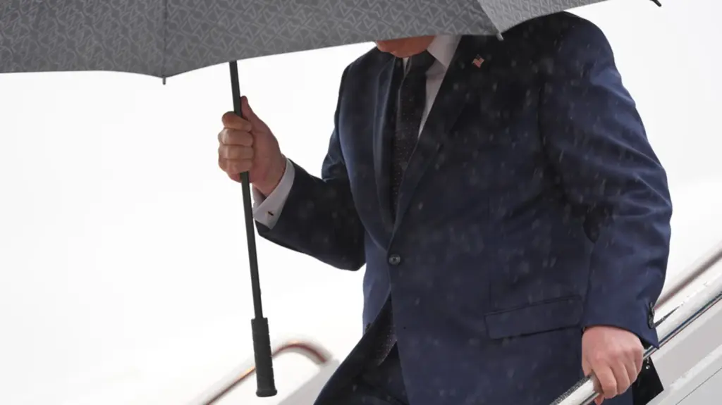 Donald Trump wearing a navy blue suit with an American flag lapel pin holds a large gray patterned umbrella while descending airplane stairs in heavy rain, gripping the handrail with his other hand. His face is obscured by the umbrella.