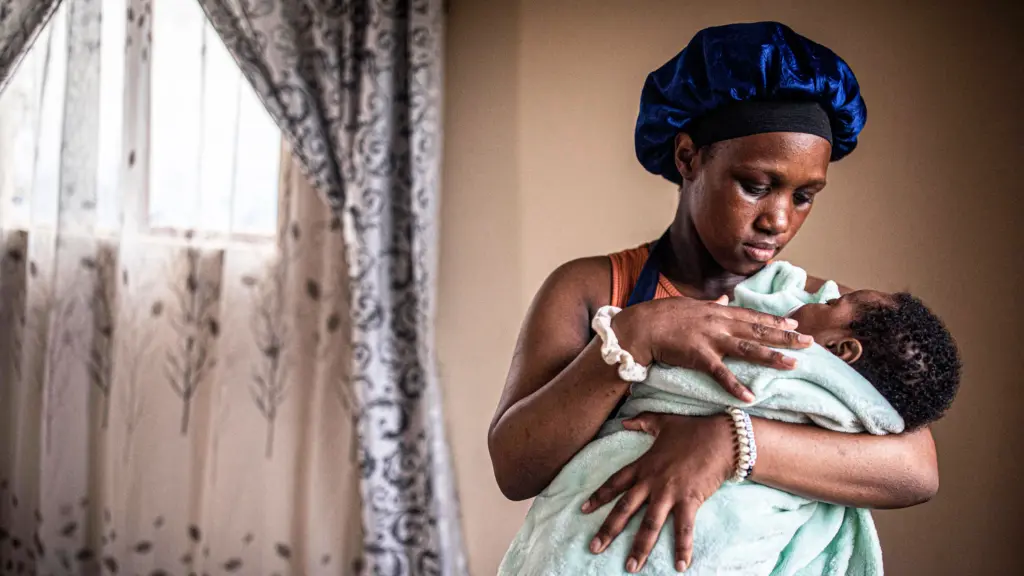 A young African American woman, with her hair in a dark blue silk bonnet, stands while holding a baby wrapped in a fuzzy, light green blanket.