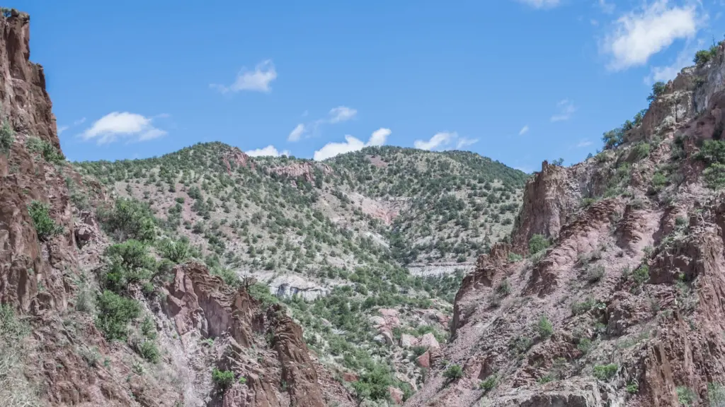 Dry, light brown mountains with sparse green shrubs against a blue sky with small clouds.