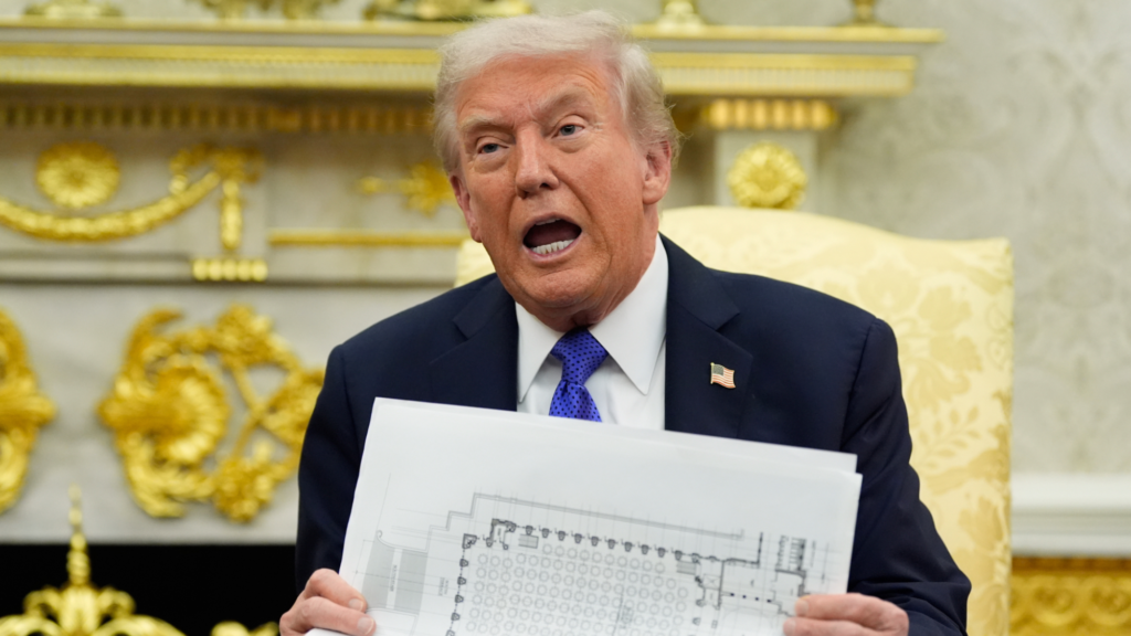 Donald Trump holds a piece of paper with a map of a table seating chart. He is sitting in a gold chair and is wearing a black suit and blue tie. He is speaking inside the Oval Office.
