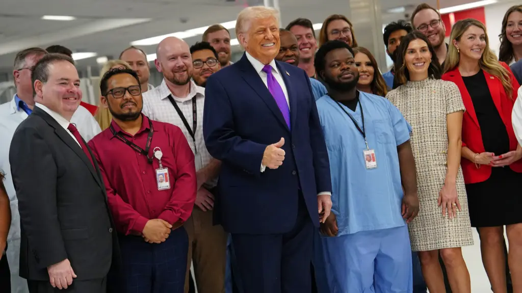 Trump stands with a group of smiling workers wearing id tags around their necks. He's wearing a purple tie and doing the thumbs up.