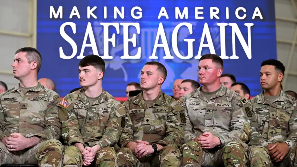 A group of soldiers in fatigues listening to someone speak; behind them is a large