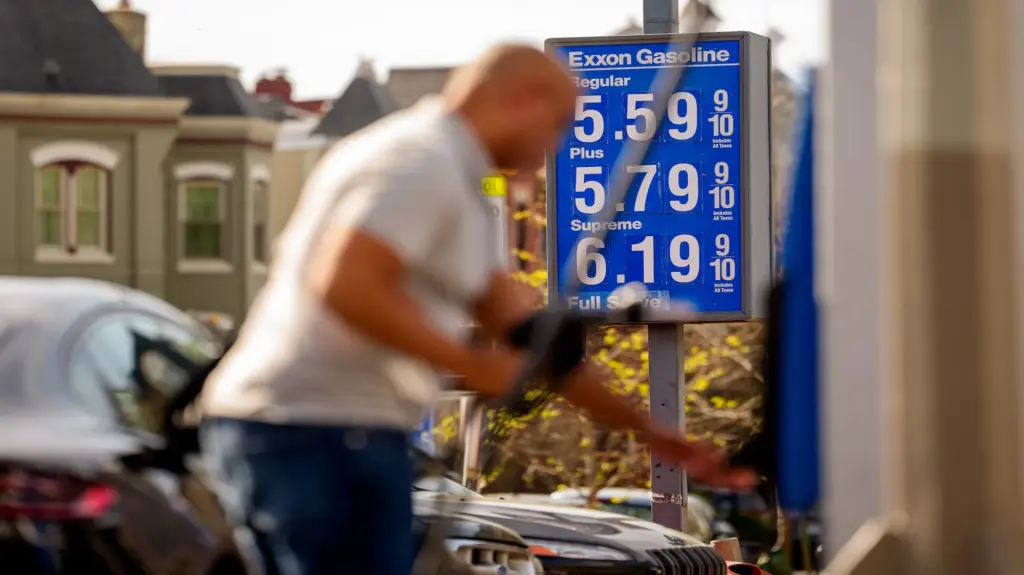 In the background, a blue Exxon gas station sign displays prices over $5 per gallon. In the foreground, an out-of-focus figure pays for gas.