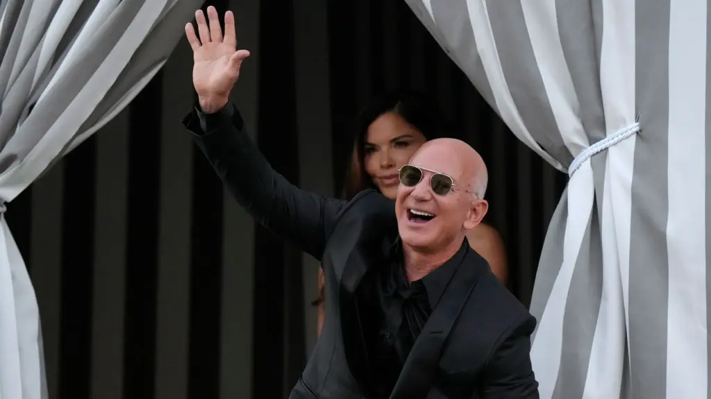 A happy looking well-dressed bald man in sunglasses waves and smiles as his new wife looks on from behind him