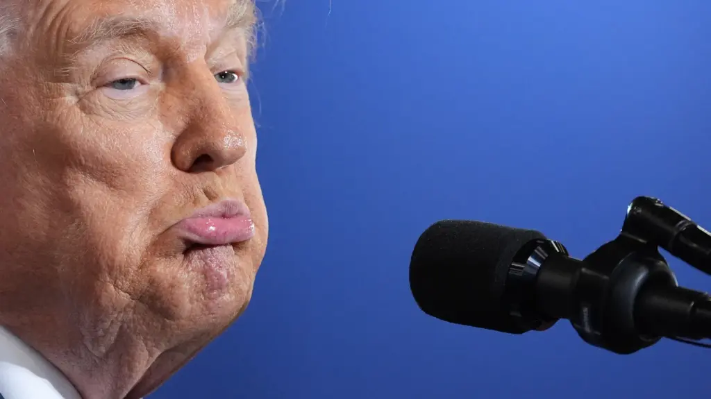 A tight close-up of Donald Trump's face as he speaks into a black podium microphone, set against a solid blue backdrop. He wears a dark tie with a white shirt collar visible.