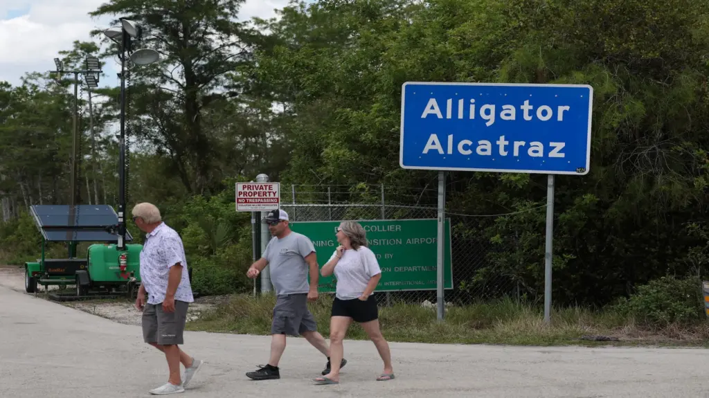 Three people walk past a wooded area and a large blue sign that reads