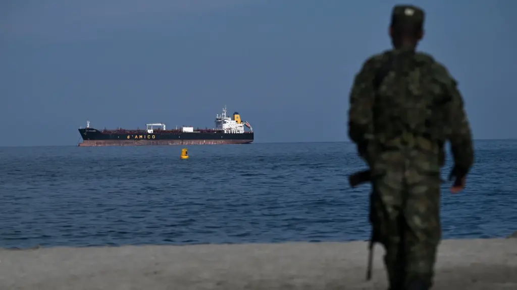 A man in camo with an rifle walks on a beach with his back to the camera. An oil tanker passes by in the water in front of him.