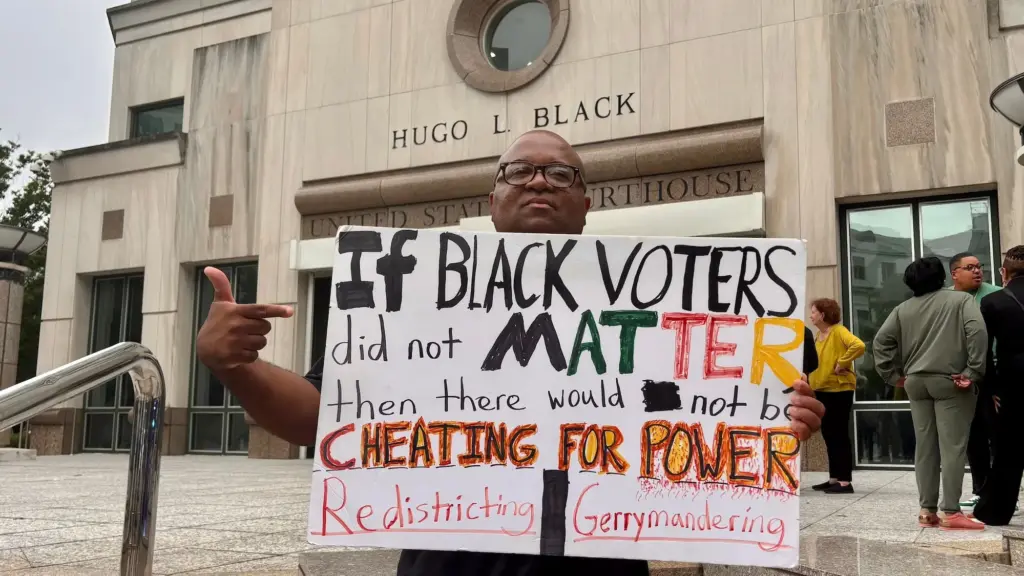 A Black man wearing glasses holds a protest sign that reads: