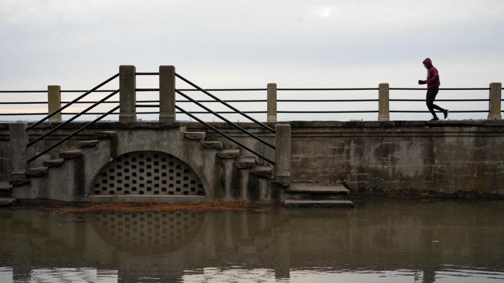 Under a cloudy sky, a jogger runs in a hoodie along a grey battery beside reflective water flooding the street.