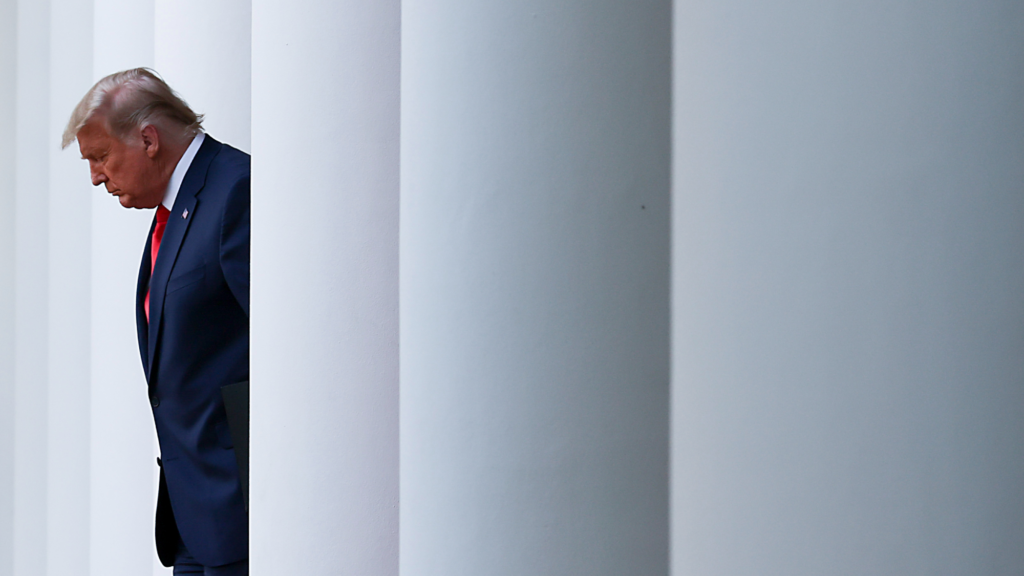 President Donald Trump is looking downward while walking through white columns outside. He is wearing a dark suit and red tie.