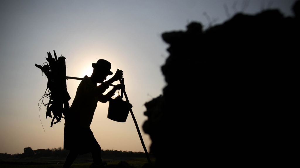 Silhouette of a man walking through a field holding a long stick and a bucket