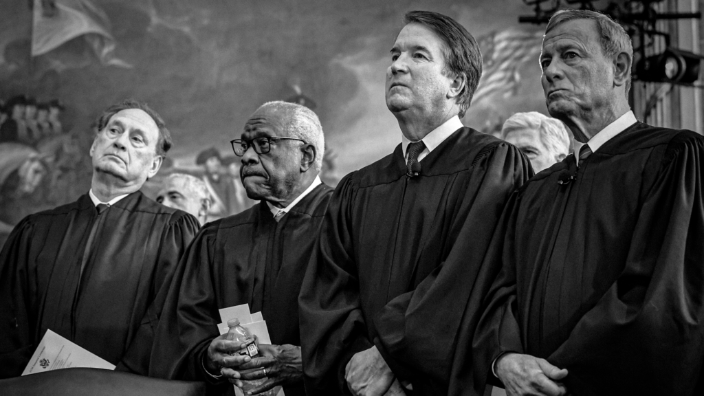A black-and-white photo of US Associate Supreme Court Justices Samuel Alito, Jr., Clarence Thomas and Brett Kavanaugh standing beside US Supreme Court Chief Justice John Roberts. They are all wearing robes and ties, looking straight ahead stoically.