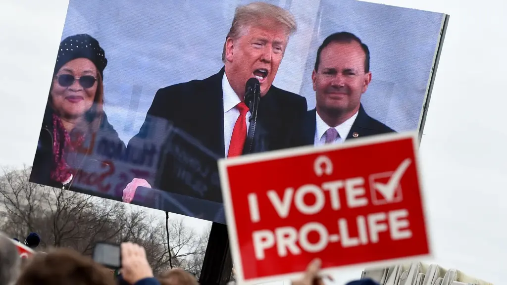 Trump is depicted speaking on a large screen in front of a crowd. One person in the crowd holds up a red sign that says