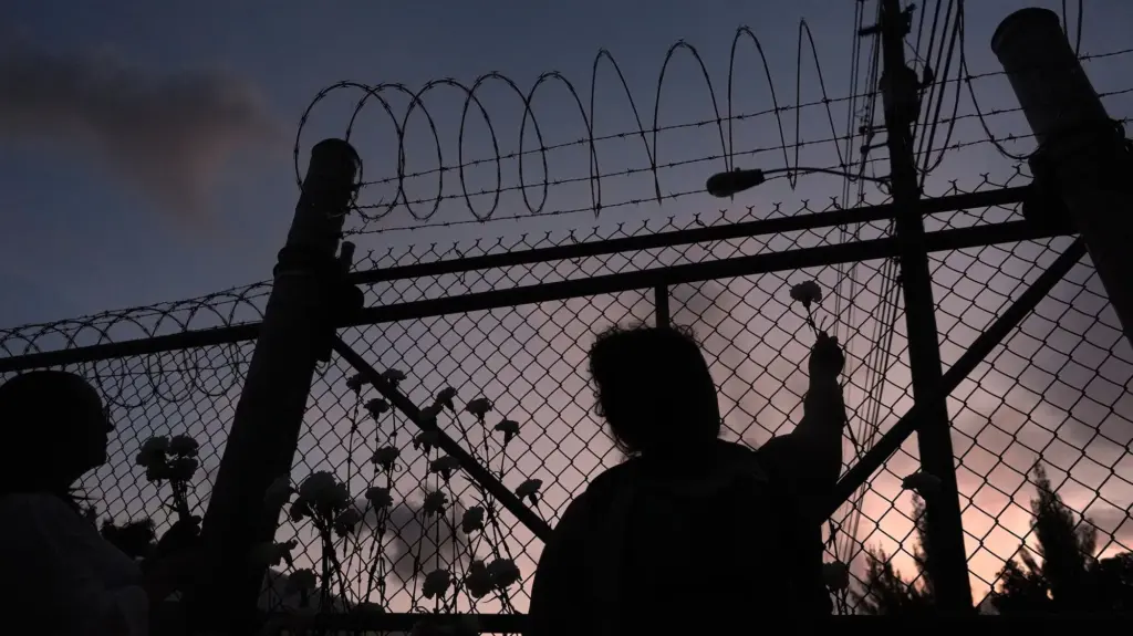 A person holds a flower outside a chain-link and barbed-wire fence