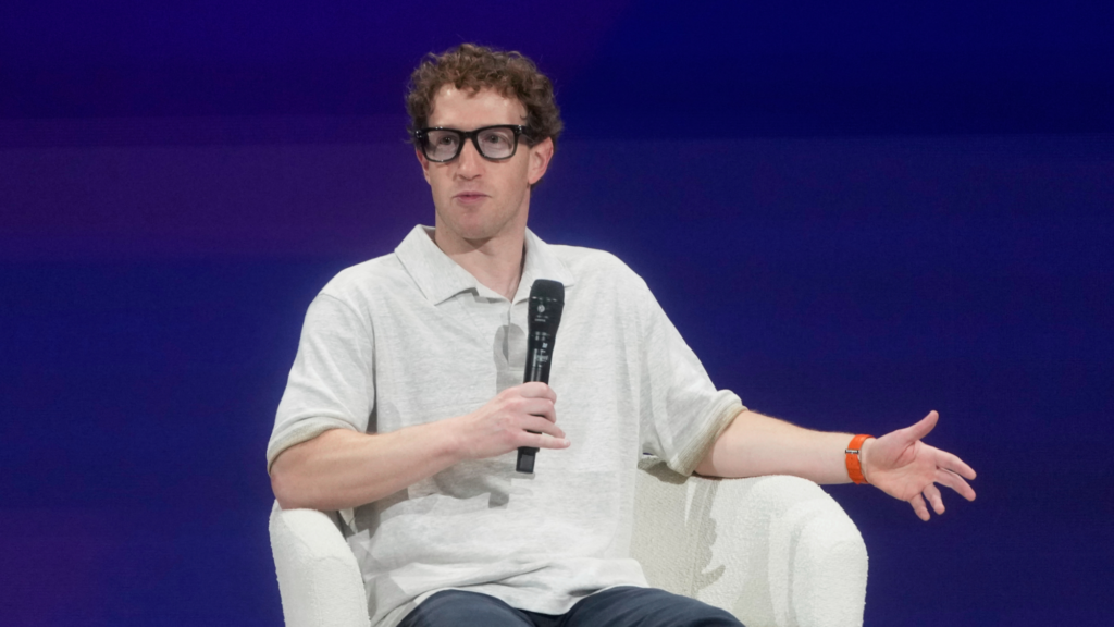 Mark Zuckerberg is wearing a white-gray collared shirt and is sitting in a white chair. He is speaking at a conference and is holding a black microphone to his mouth with his right hand. He is wearing thick-rimmed black glasses.