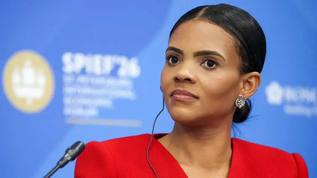Candace Owens, a 37 year old African American woman, before a microphone wearing a red jacket. A blue backdrop displays a conference logo.