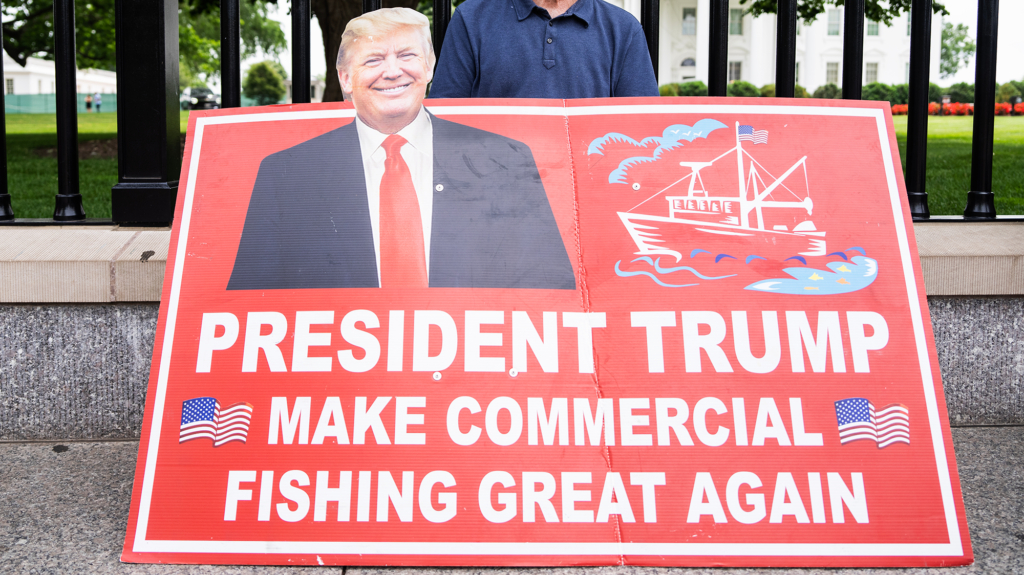 Red sign with white lettering has picture of Donald Trump in suit and red tie. The sign reads 