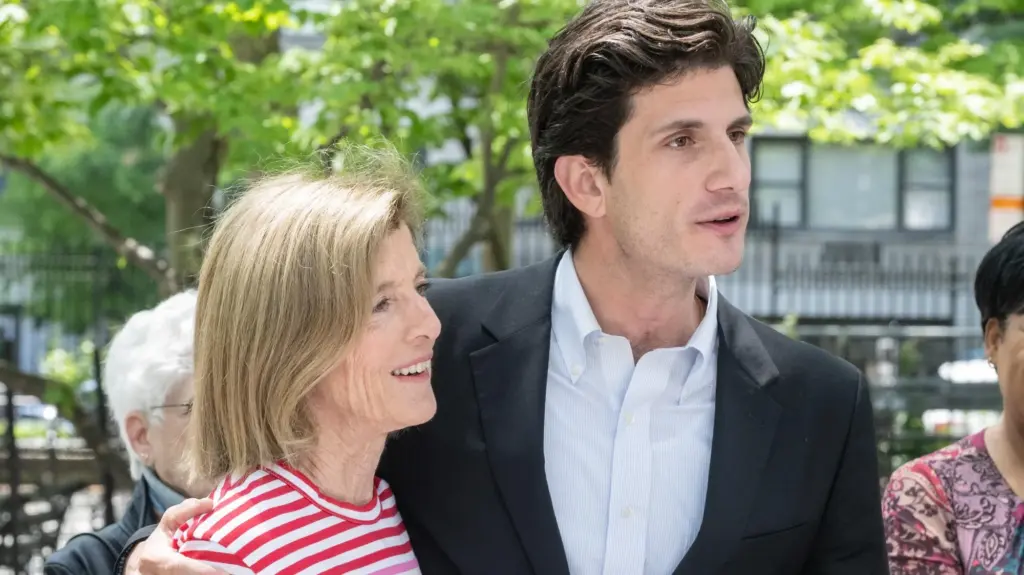 Schlossberg, a 33 year old man in a suit with no tie, has his arm around Kennedy, a 68 year old woman in a red-and-white striped shirt.
