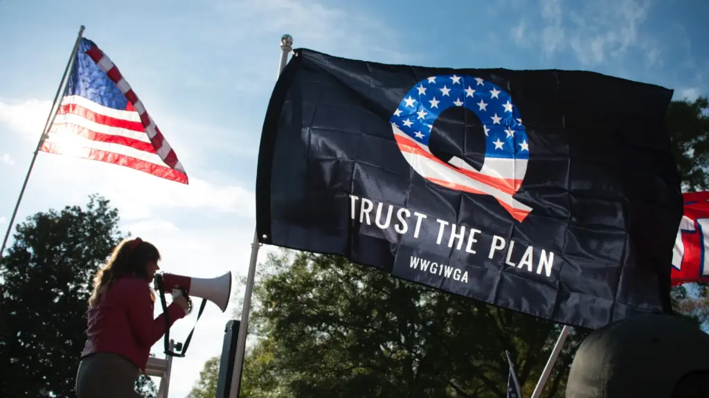 A smaller US flag waves to the left of a large black flag with a Q on it that reads