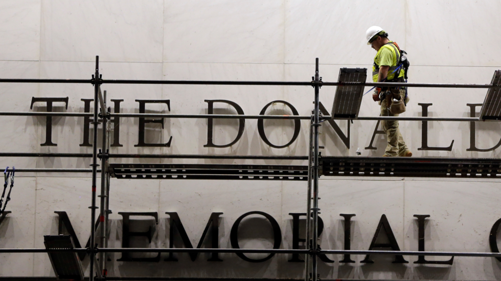 One worker, wearing a white helmet, is walking on scaffolding to remove part of a sign in front of the Kennedy Center.