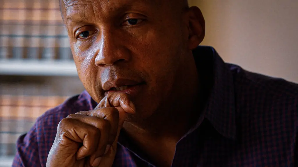 A close-up photograph of a middle-aged Black man who is staring downward pensively as he holds his right hand up to his chin. The man has a shaved head and wears a long-sleeved shirt with a blue-and-purple checkered pattern. On his wrist is a bracelet with red beads. Behind him, out of focus, is a wall of books.
