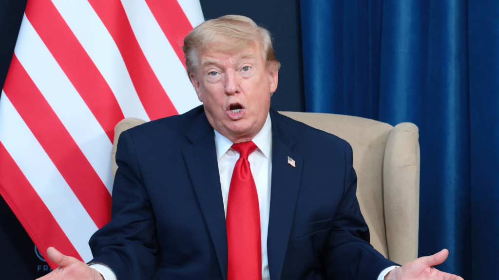 Donald Trump speaks while sitting in a brown chair. He is wearing a suit and both of his hands are open in front of him in a nonchalant way.