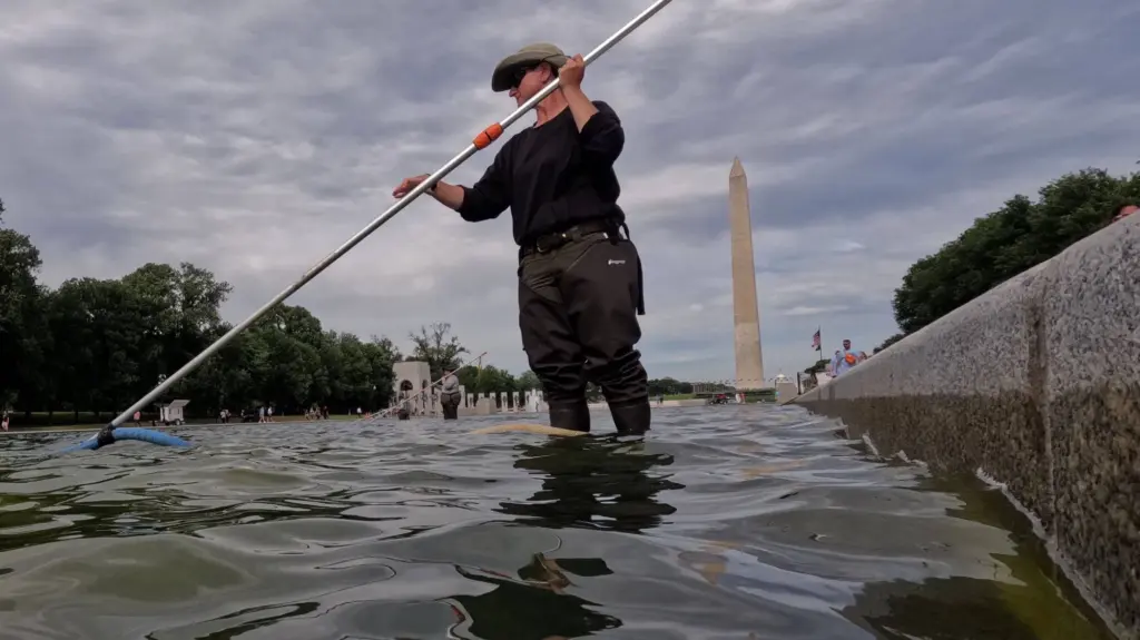 A man wearing thigh-high waders maneuvers a tube mounted on a metal pole while standing calf-deep in the murky water of DC's reflecting pool. The Washington monument can be seen behind him.
