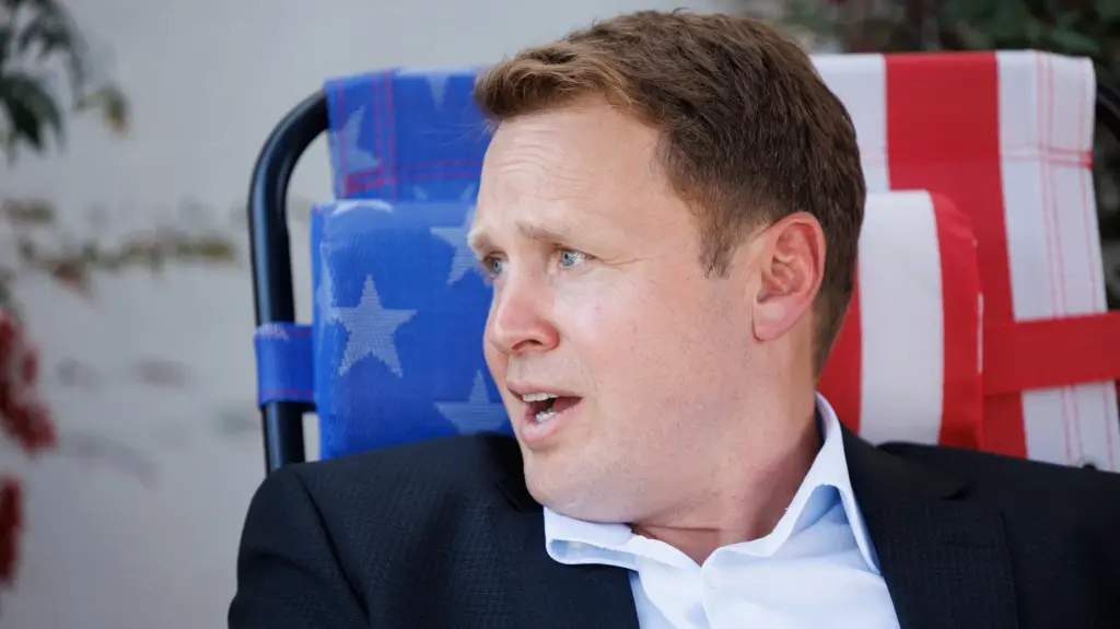 A white man in a suit coat with no tie sits on an American flag patterned folding lounge chair.