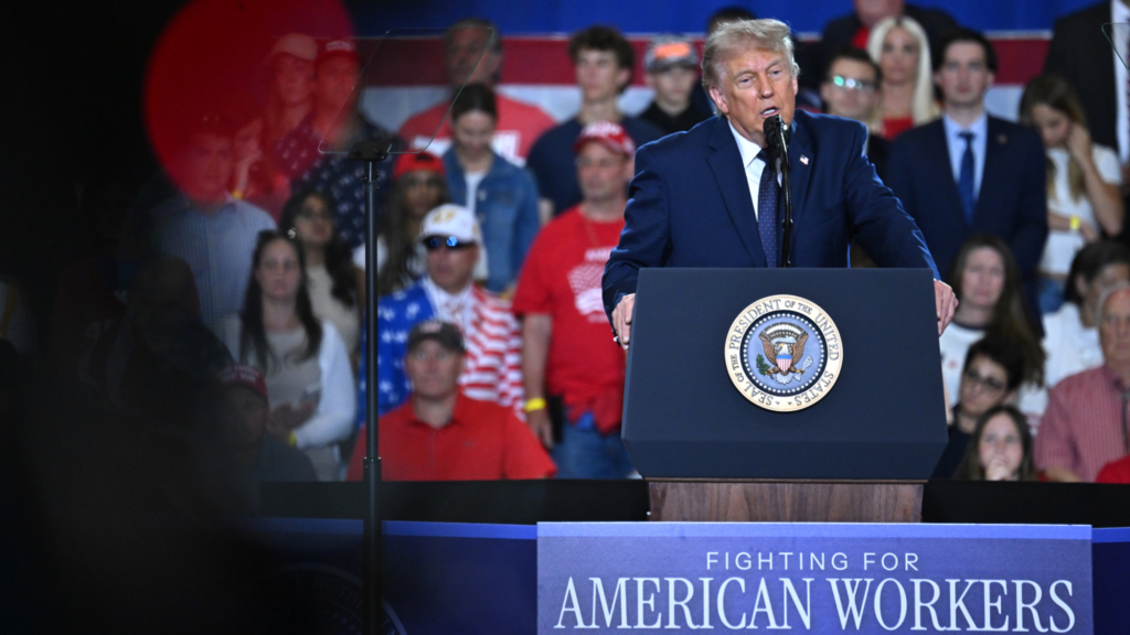 President Trump is wearing a dark blue suit and blue tie. He is delivering a speech and speaking into a microphone on a podium. Behind him are over a dozen audience members at a political rally. A sign underneath the president reads: