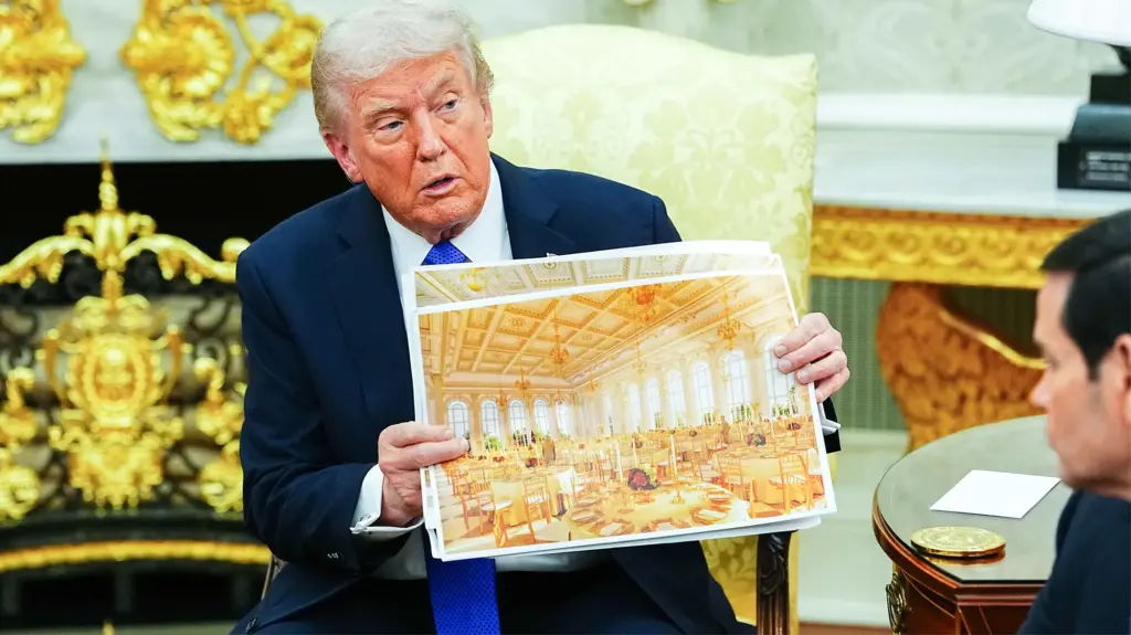 President Donald J. Trump is sitting in the Oval Office and presenting a large architectural rendering of a golden ballroom to other people in the room.