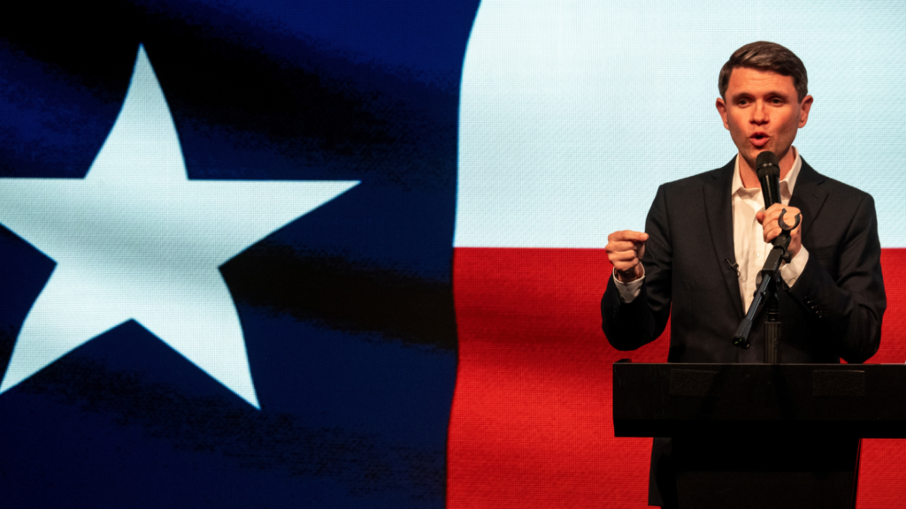 James Talarico is giving a speech. He is holding a microphone in his left hand and is standing behind a podium. A projected image of the Texas state flag is behind him.