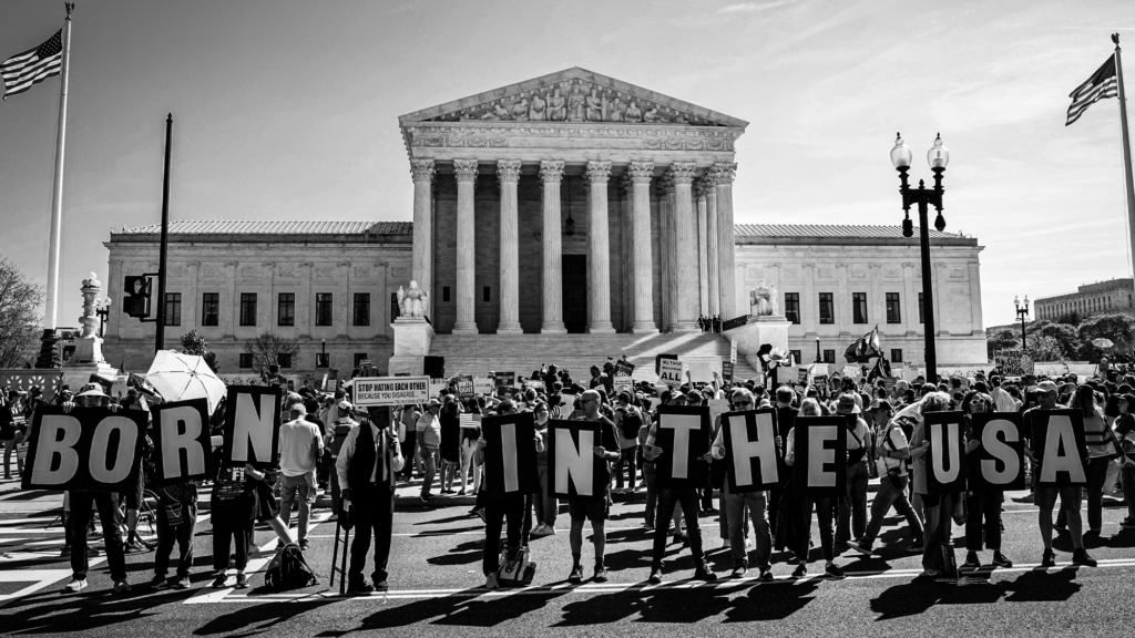 A black-and-white photo of demonstrators rallying in support of birthright citizenship outside the US Supreme Court. Individuals in front of the building's steps hold placards that each have one letter. Together, their signs spell