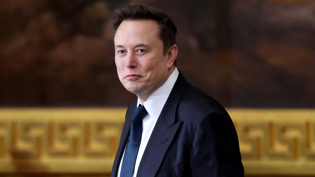 Elon Musk, a white dude with brown hair, wearing a jacket and blue tie and looking smug
