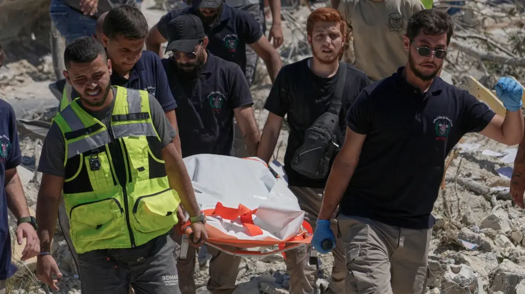 Five men, with one wearing a yellow high visibility vest, carry an orange stretcher covered by a white tarp across a field of rubble.