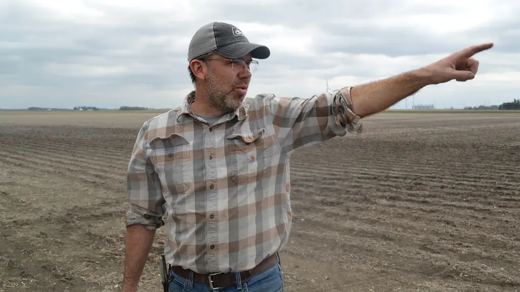 A white man in cap and plaid shirt stands in a fallow field on a cloudy day, pointing to something in the distance.