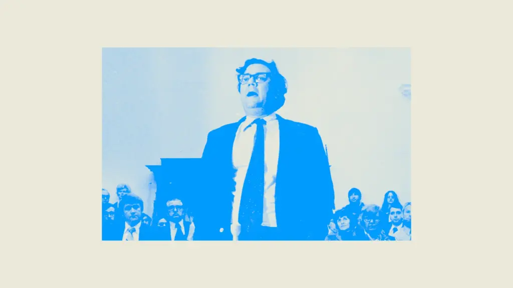 A high-contrast duotone photograph in shades of bright blue and cream, showing a person with dark hair and glasses wearing a dark suit jacket, white shirt, and a tie. The person stands in the center foreground, facing forward with an open mouth. In the lower background, several other indistinct individuals are seated facing forward.