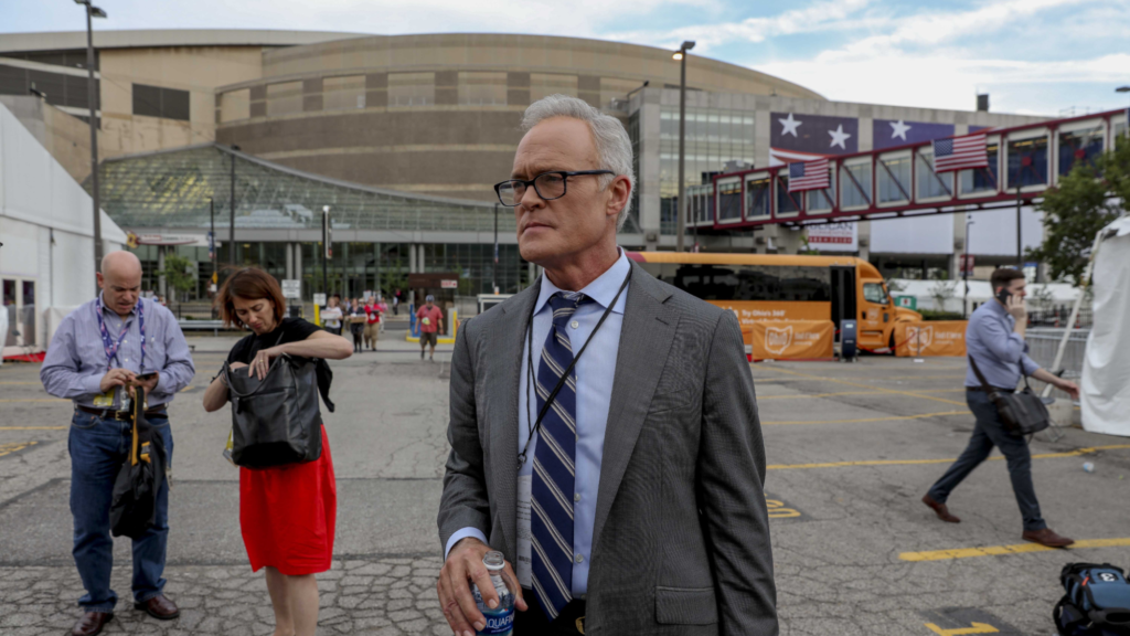 Scott Pelley, dressed in a gray suit and holding a water bottle in his right hand, stands outside a stadium. Several people are in the background working.