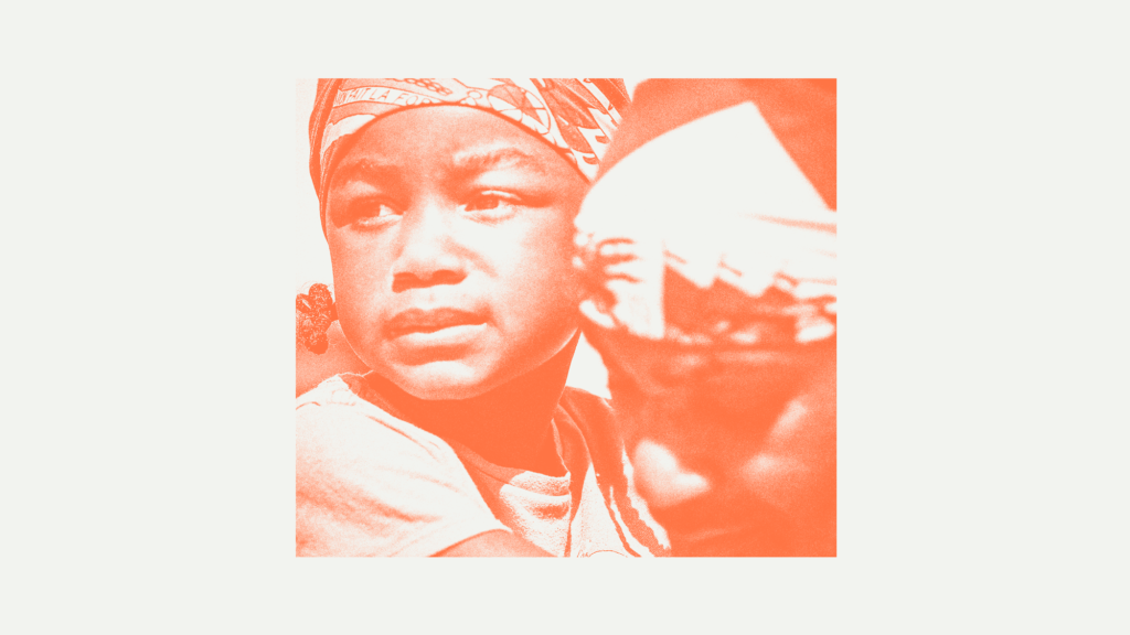 A close-up photo of a little black child holding onto the neck of her father. Both are wearing Haitian headscarves.