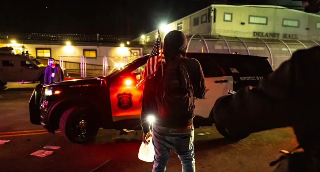A lone protester, seen from behind, stands at night in front of Delaney Hall — a detention facility in Newark, New Jersey. The figure, wearing a dark hoodie and jeans, holds a small American flag over one shoulder and a glowing flashlight or lantern in their other hand. They face a Newark Metro Precinct police cruiser (unit 212) with its brake lights casting a red glow across the scene. A second black-and-white emergency vehicle marked