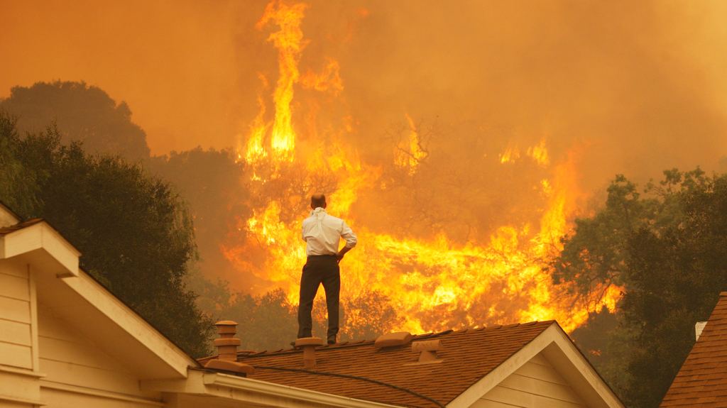 Man in white shirt and black pants stands on top of white house looking at a fire burning behind a house.