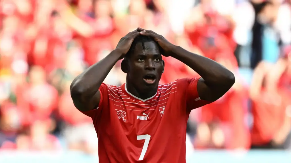 Breel Embolo, a dark-complexioned man in a red number 7 soccer jersey, presses his hands to the top of his head and looks dismayed.