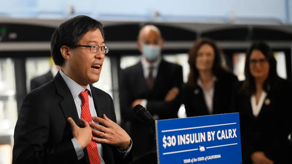 A man with glasses and a suit with a red tie speaks in front of a sign that says $30 insulin on Cal Rx