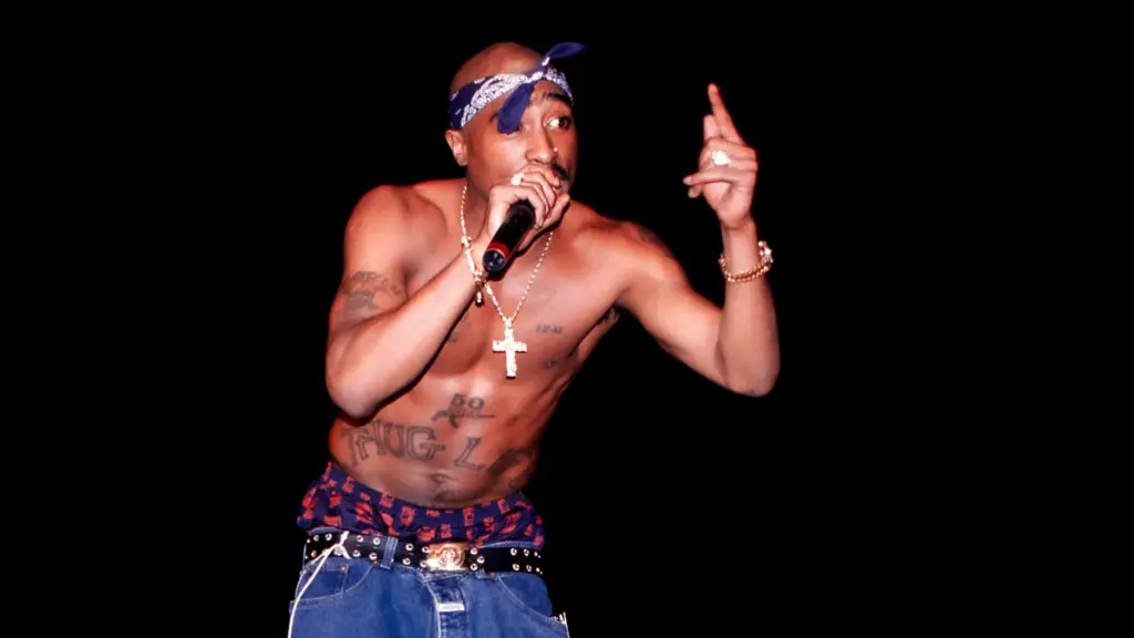 A young Black man holds a microphone to his mouth with his right hand as he gestures with his left hand. Shirtless, he wears baggy jeans over blue-and-red boxers that peek out, a blue bandanna tied around his head, and a cross with a gold chain that dangles from his neck. Among the tattoos on his arms and torso, the most prominent one, just above his belly button, reads “THUG LIFE.”