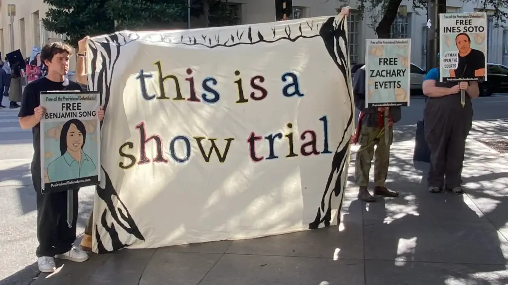 Three people hold signs reading free Benjamin Song, free Zachary Evetts and free Ines Soto around a large banner reading this is a show trial on a street corner